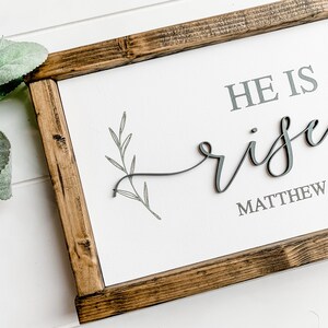 Op de afbeelding: Een houten ingelijst bord met de tekst "HE IS risen" in grijze letters met een klein grijs takontwerp. De tekst "MATTHEW" is ook in grijze letters opgenomen.