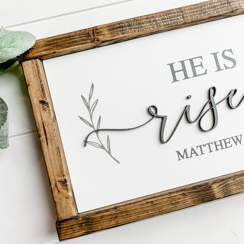 Op de afbeelding: Een houten ingelijst bord met de tekst "HE IS risen" in grijze letters met een klein grijs takontwerp. De tekst "MATTHEW" is ook in grijze letters opgenomen.