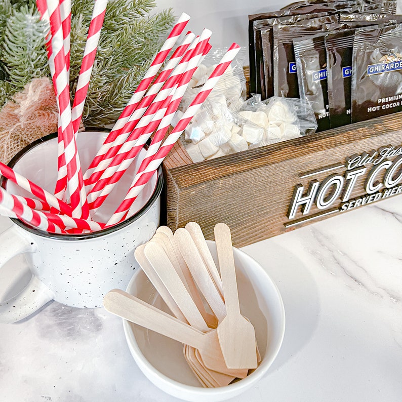 May include: A white ceramic mug filled with red and white striped paper straws. A wooden box with the text "Old Fashioned Hot Cocoa Served Here" holds white marshmallows and individual packets of hot cocoa mix. A white bowl with wooden spoons is on the counter.