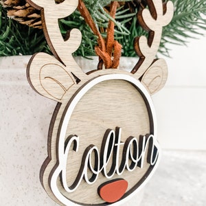 May include: Wooden reindeer ornament with a white outline and the name "Colton" written in black cursive. The ornament has a red nose and a brown suede cord.