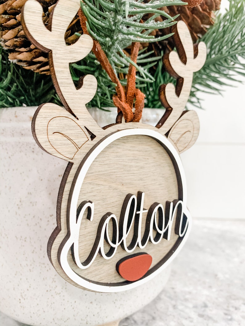 May include: Wooden reindeer ornament with a white outline and the name "Colton" written in black cursive. The ornament has a red nose and a brown suede cord.
