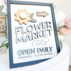 May include: A wooden sign with a black frame and white background. The sign says "Flower Market" in gray raised letters. A yellow flower with a white center is in the upper left corner. An arrow points to the right. The sign also says "Open Daily" in gray raised letters and "Peonies, Tulips, Roses" in smaller gray letters.