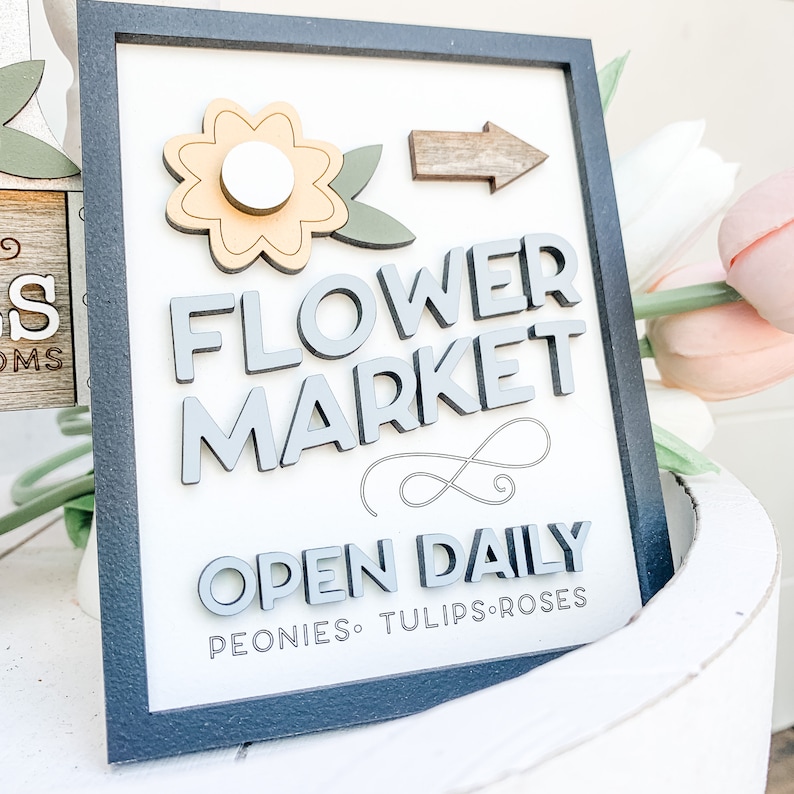 May include: A wooden sign with a black frame and white background. The sign says "Flower Market" in gray raised letters. A yellow flower with a white center is in the upper left corner. An arrow points to the right. The sign also says "Open Daily" in gray raised letters and "Peonies, Tulips, Roses" in smaller gray letters.