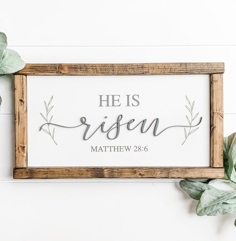 Op de afbeelding: Een wit houten bord met een bruin houten frame. Het bord zegt "Hij is opgestaan" in cursief schrift met een grijze achtergrond. Het bord bevat ook de Bijbelverwijzing "Matthe&uuml;s 28:6".