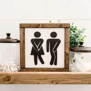 Puede incluir: Un letrero enmarcado en madera con fondo blanco y siluetas negras de un hombre y una mujer en un icono de estilo de ba&ntilde;o. El letrero est&aacute; sentado en un estante de madera.