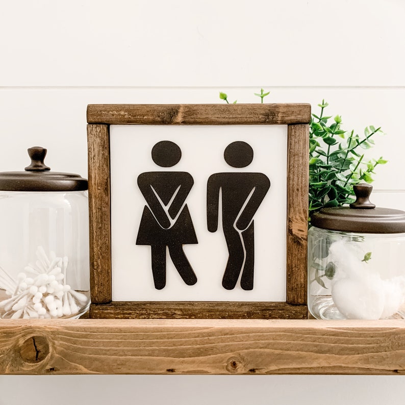 Puede incluir: Un letrero enmarcado en madera con fondo blanco y siluetas negras de un hombre y una mujer en un icono de estilo de ba&ntilde;o. El letrero est&aacute; sentado en un estante de madera.