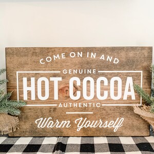 May include: A wooden sign with the text "Come on in and Genuine Hot Cocoa Authentic Warm Yourself" in white lettering on a brown background.