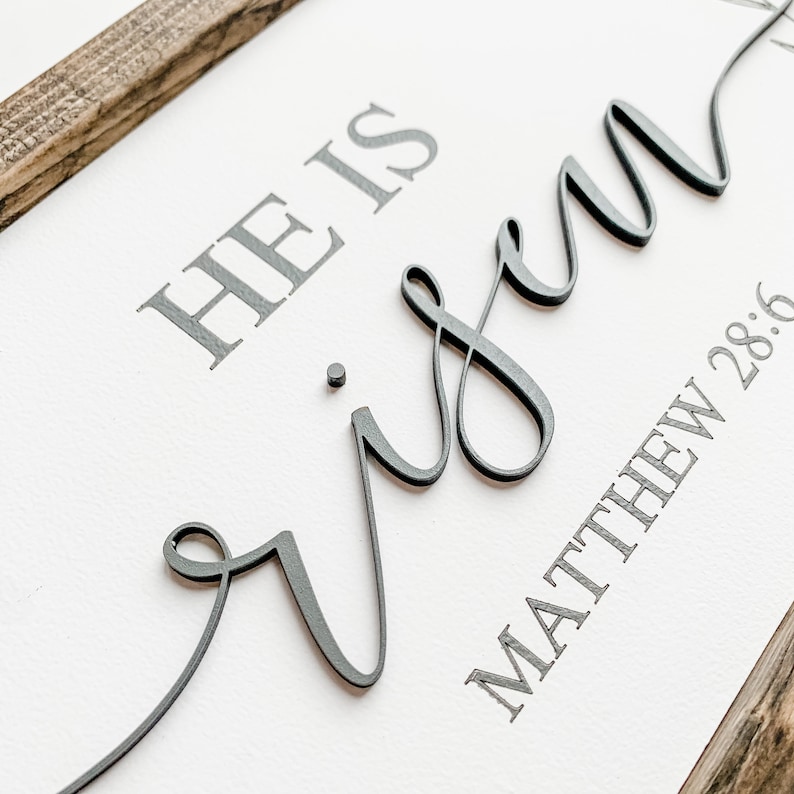 Op de afbeelding: Een wit houten bord met de tekst "HE IS risen" in grijze letters. Het woord "risen" is in een cursief lettertype en steekt uit boven het oppervlak van het bord. De onderkant van het bord heeft de tekst "MATTHEW 28:6" in grijze letters.