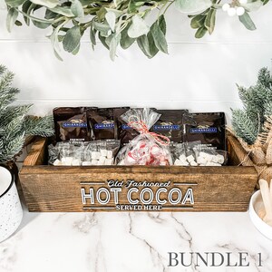 May include: A wooden box with the text "Old Fashioned HOT COCOA SERVED HERE" on the front. The box contains several small bags of hot cocoa mix, marshmallows, and candy canes.