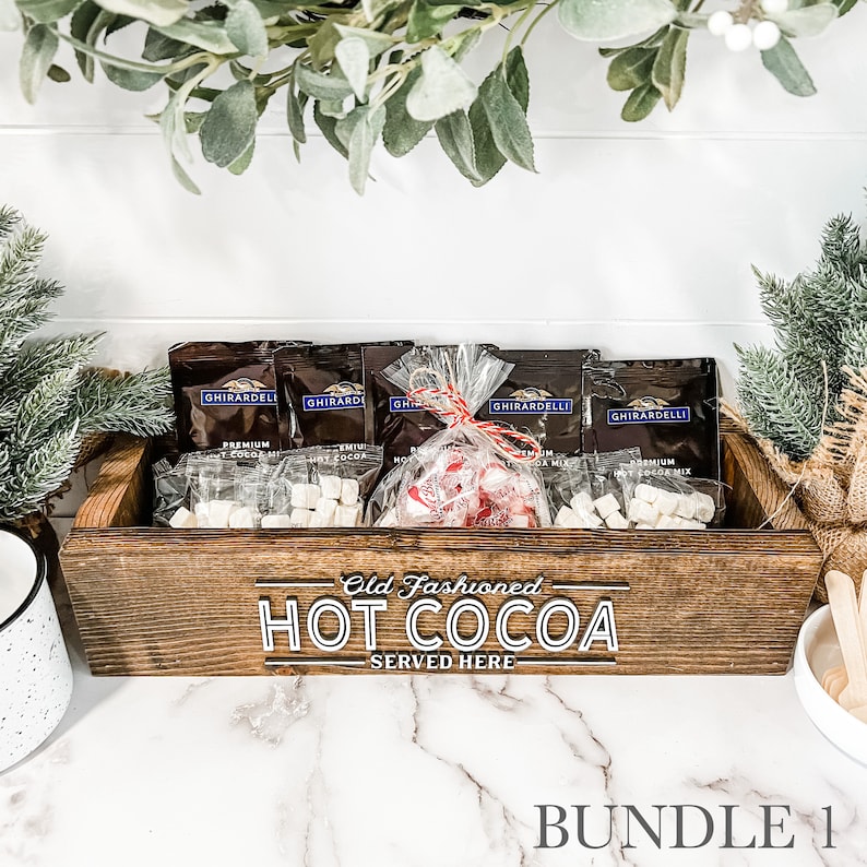 May include: A wooden box with the text "Old Fashioned HOT COCOA SERVED HERE" on the front. The box contains several small bags of hot cocoa mix, marshmallows, and candy canes.