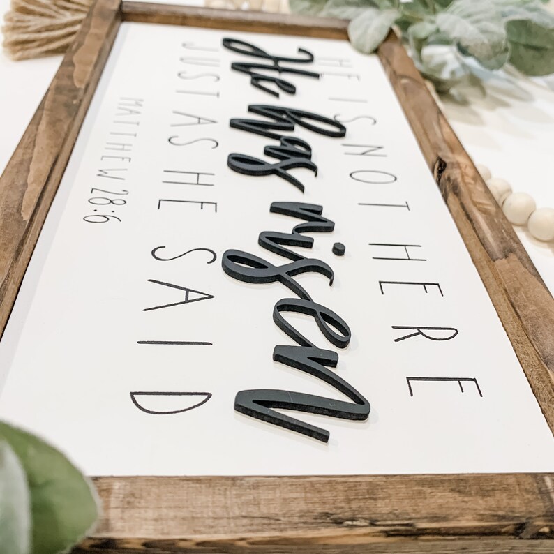 May include: A white wooden sign with a brown frame. The sign says "He is not here, he has risen, just as he said" in black text. The words "he has risen" are in a larger font and are raised from the surface of the sign. The sign also includes the scripture reference "Matthew 28:6".