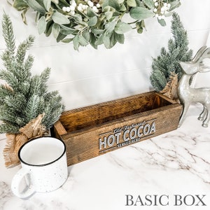 May include: A rustic wooden box with the text "Old Fashioned Hot Cocoa Served Here" in white lettering. The box is sitting on a white marble surface with a white mug and a silver reindeer figurine.
