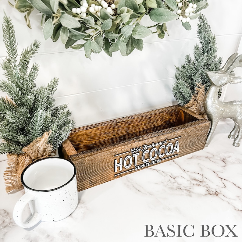 May include: A rustic wooden box with the text "Old Fashioned Hot Cocoa Served Here" in white lettering. The box is sitting on a white marble surface with a white mug and a silver reindeer figurine.