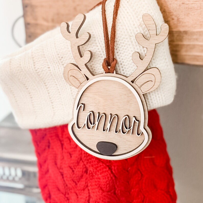 May include: A wooden reindeer ornament with the name "Connor" engraved on it. The ornament is hanging from a red and white stocking.