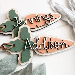 K&ouml;nnte beinhalten: Zwei h&ouml;lzerne Namensschilder in Karottenform mit gr&uuml;nen Blattwerk und braunem Lederband. Auf dem einen Schild steht der Name "Jennings" und auf dem anderen "Addison".