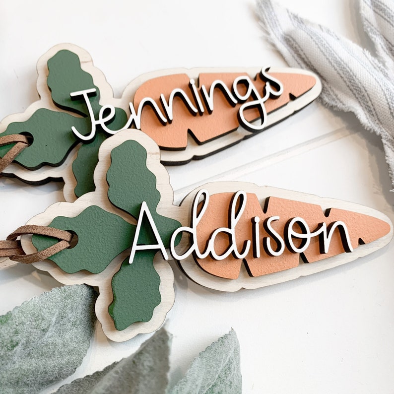 K&ouml;nnte beinhalten: Zwei h&ouml;lzerne Namensschilder in Karottenform mit gr&uuml;nen Blattwerk und braunem Lederband. Auf dem einen Schild steht der Name "Jennings" und auf dem anderen "Addison".