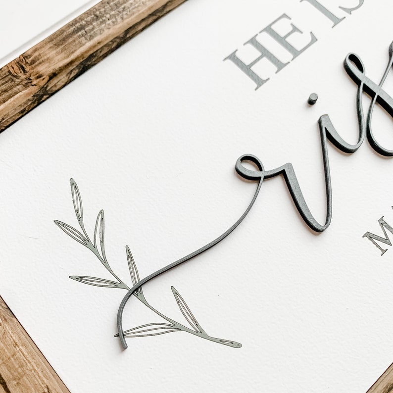 Op de afbeelding: Een wit houten bord met de tekst "He is risen" in grijze letters. Het bord heeft een zwarte, cursieve "risen" met een klein, grijs, bladerig takje in de linkerbenedenhoek.