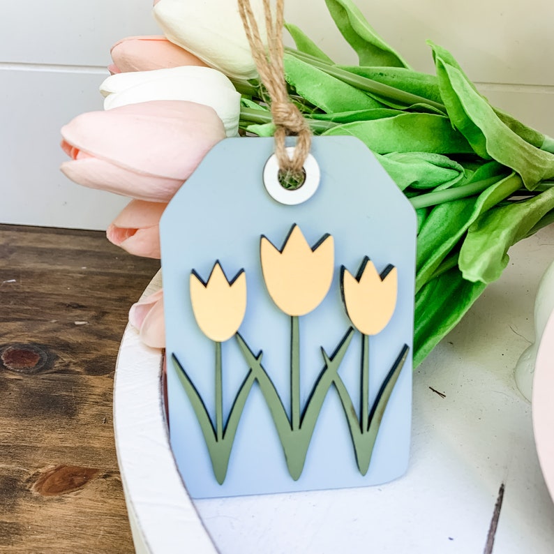 May include: A light blue wooden tag with three yellow tulips with green stems and leaves. The tag has a hole at the top and is tied with twine.