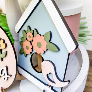 May include: A light blue wooden birdhouse with a white roof and a pink bird cutout. The birdhouse is decorated with green leaves and pink flowers.