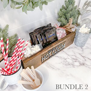 May include: A wooden box with the text "Old Fashioned Hot Cocoa Served Here" on the front. The box is filled with individual packets of hot cocoa mix, marshmallows, and red and white striped straws.