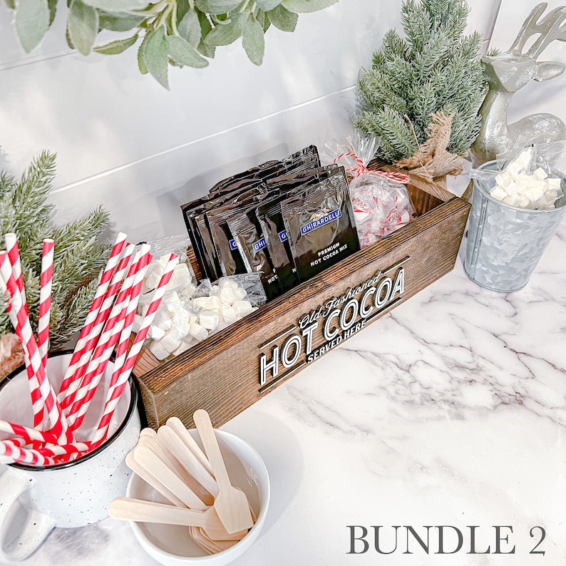 May include: A wooden box with the text "Old Fashioned Hot Cocoa Served Here" on the front. The box is filled with individual packets of hot cocoa mix, marshmallows, and red and white striped straws.