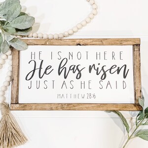May include: A wooden sign with a white background and a brown frame. The sign has black text that reads "HE IS NOT HERE He has risen JUST AS HE SAID MATTHEW 28:6".