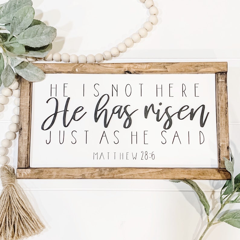 May include: A wooden sign with a white background and a brown frame. The sign has black text that reads "HE IS NOT HERE He has risen JUST AS HE SAID MATTHEW 28:6".