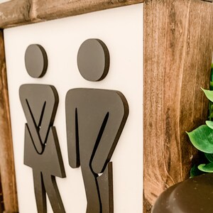 Puede incluir: Un letrero de madera con fondo blanco y figuras de silueta negra de un hombre y una mujer. El letrero tiene un marco de madera marr&oacute;n.