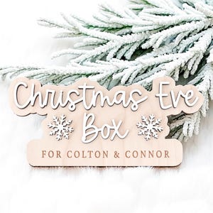 May include: A wooden Christmas Eve Box with white lettering and snowflake accents. The text reads "Christmas Eve Box" and "FOR COLTON & CONNOR". The box is light brown and sits in front of a frosted evergreen branch.