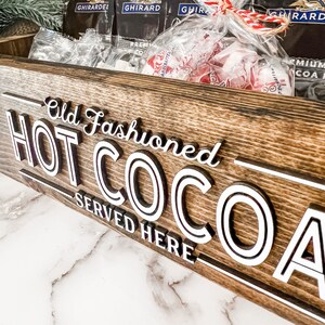 May include: A wooden sign with the text "Old Fashioned HOT COCOA SERVED HERE" in white lettering. The sign is brown and has a rustic look.