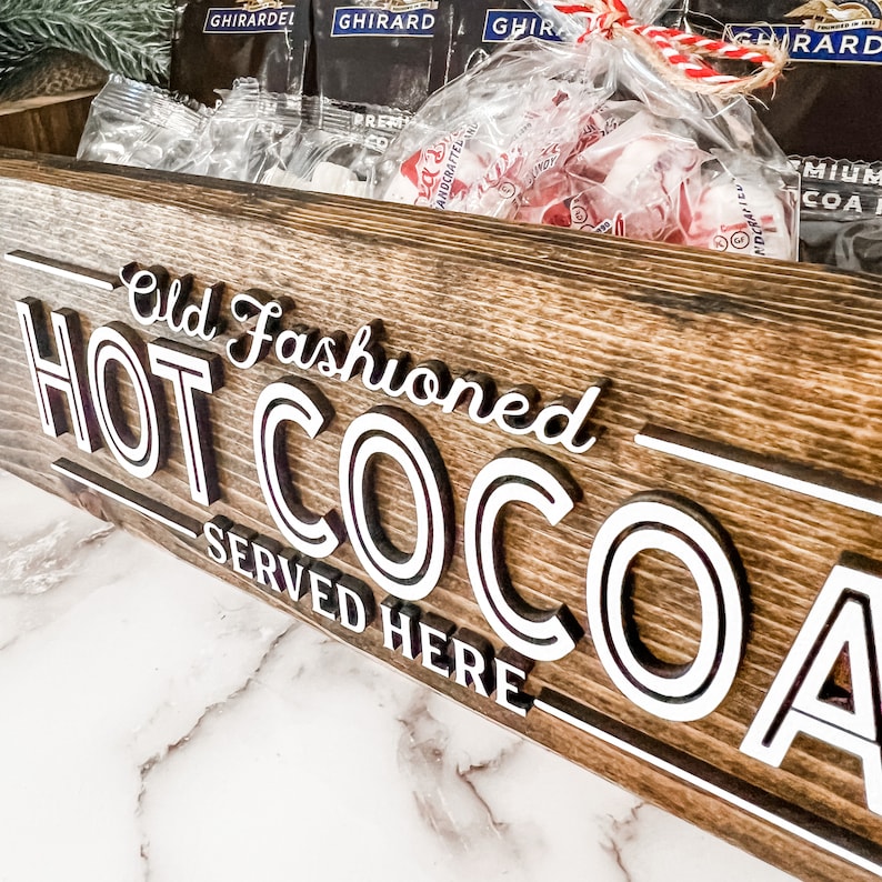 May include: A wooden sign with the text "Old Fashioned HOT COCOA SERVED HERE" in white lettering. The sign is brown and has a rustic look.