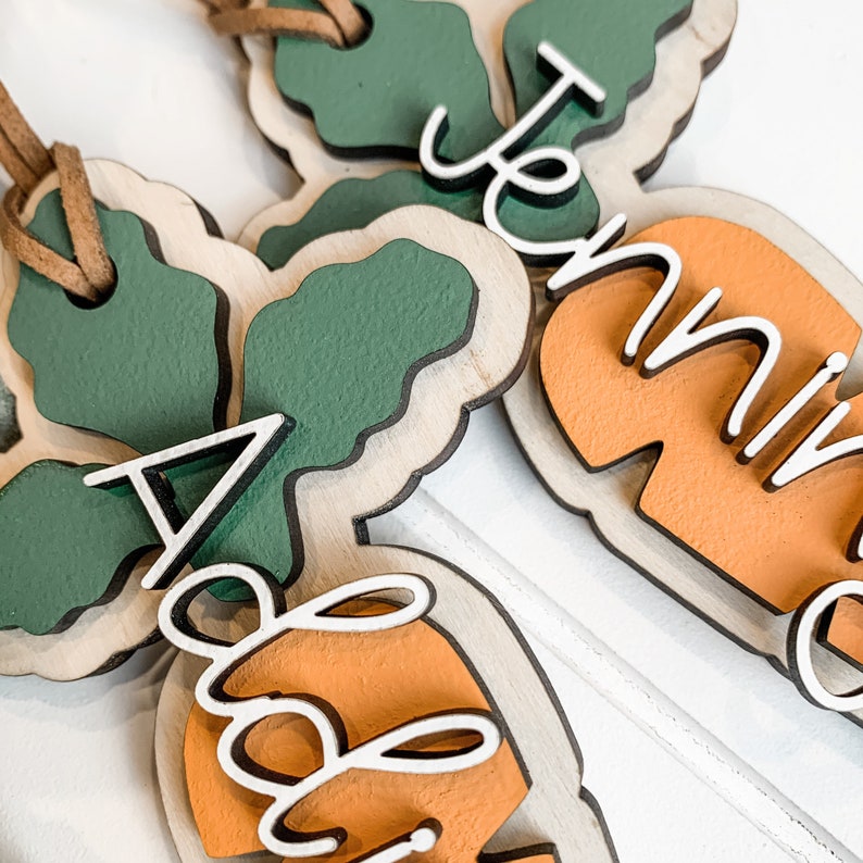 K&ouml;nnte beinhalten: Zwei h&ouml;lzerne Karotten-Anh&auml;nger mit gr&uuml;nen Spitzen und orangefarbenen Karottenk&ouml;rpern. Die Karotten sind mit wei&szlig;en Buchstaben verziert, eine sagt "Abby" und die andere sagt "Jenna".