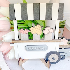 May include: A white wooden flower stand with a black and white striped awning. The stand has a sign that says "Fresh Flowers" and features three different types of flowers: pink, yellow, and orange. The stand has wooden wheels and a brown wooden base.