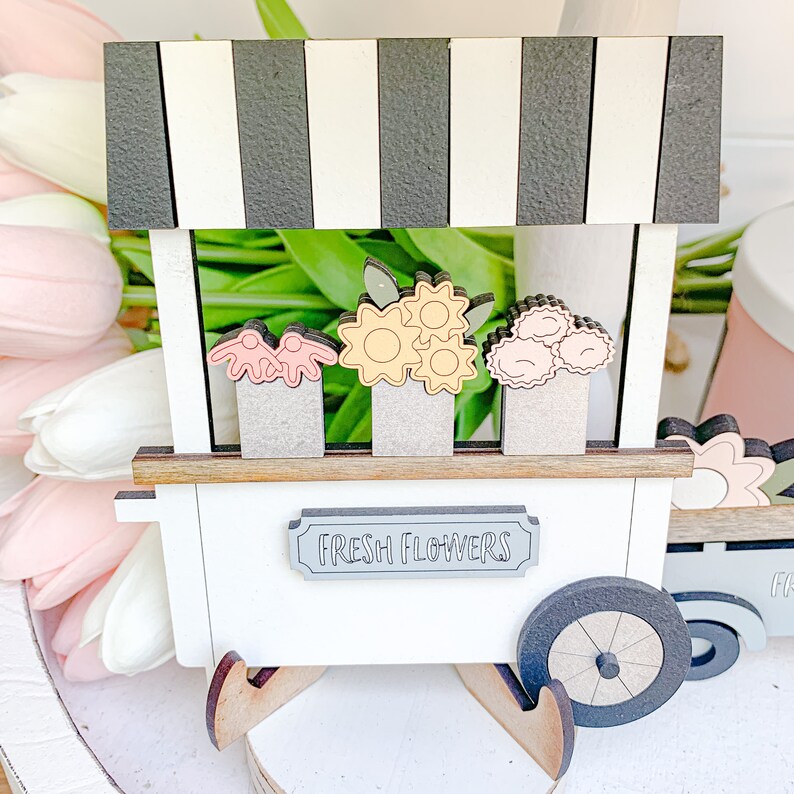 May include: A white wooden flower stand with a black and white striped awning. The stand has a sign that says "Fresh Flowers" and features three different types of flowers: pink, yellow, and orange. The stand has wooden wheels and a brown wooden base.