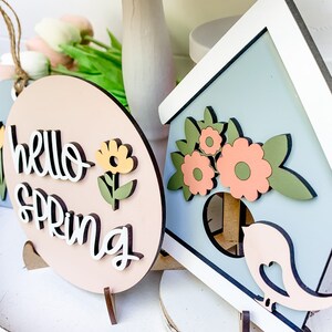 May include: Two wooden signs with a spring theme. One sign is pink and says "Hello Spring" with a yellow flower. The other sign is blue and has a birdhouse with flowers and a bird.