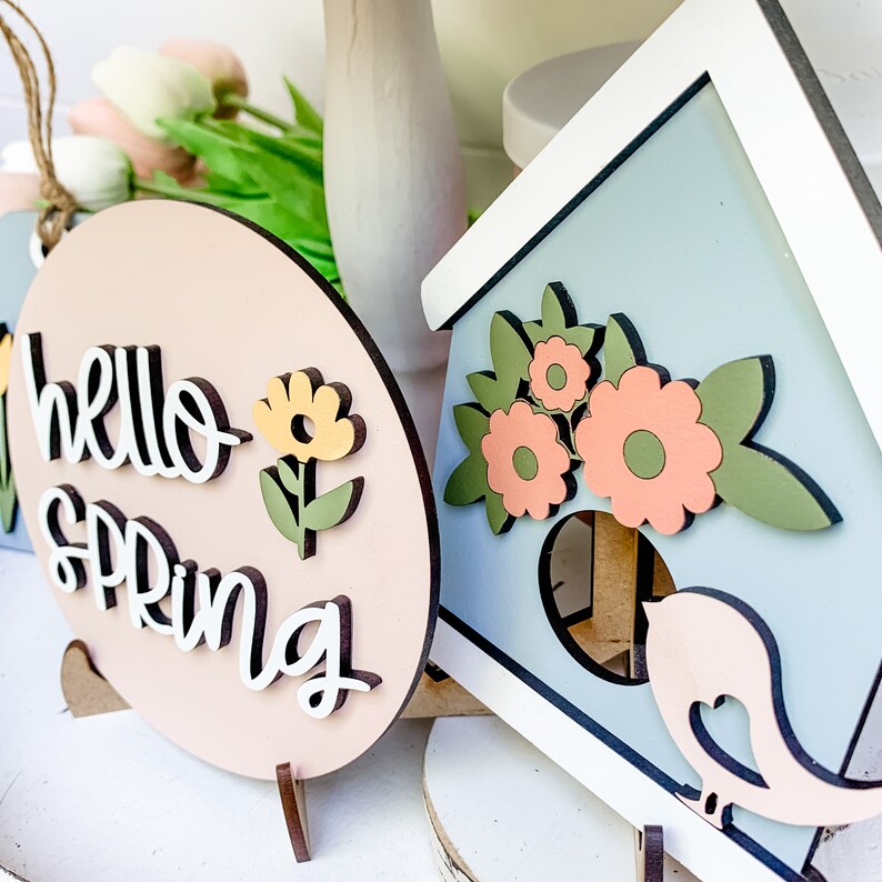 May include: Two wooden signs with a spring theme. One sign is pink and says "Hello Spring" with a yellow flower. The other sign is blue and has a birdhouse with flowers and a bird.