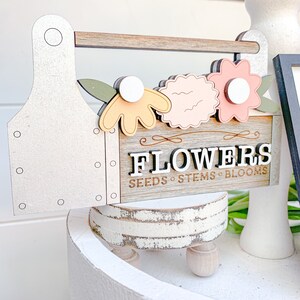 May include: A wooden toolbox sign with the word "FLOWERS" in white letters with black outlines. The sign also says "SEEDS - STEMS - BLOOMS" in smaller letters. The toolbox is decorated with three flowers in shades of pink, yellow, and green.