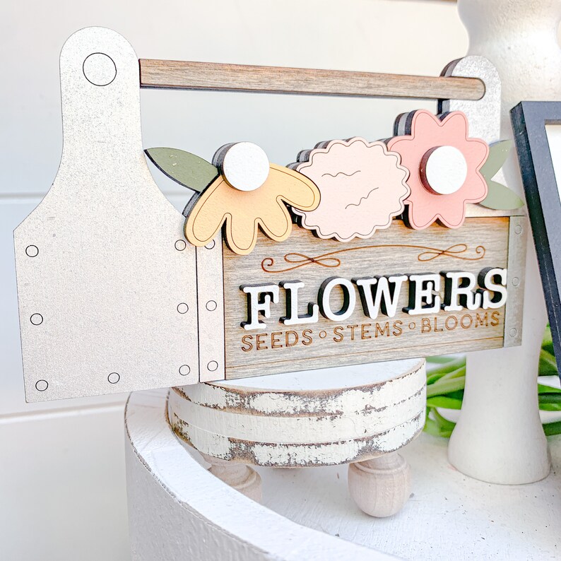 May include: A wooden toolbox sign with the word "FLOWERS" in white letters with black outlines. The sign also says "SEEDS - STEMS - BLOOMS" in smaller letters. The toolbox is decorated with three flowers in shades of pink, yellow, and green.