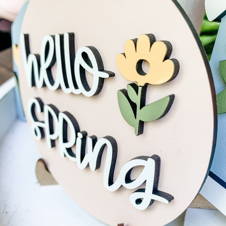 May include: A pink round wooden sign with the words "hello spring" in black and white lettering. A yellow flower with green leaves is on the right side of the sign.