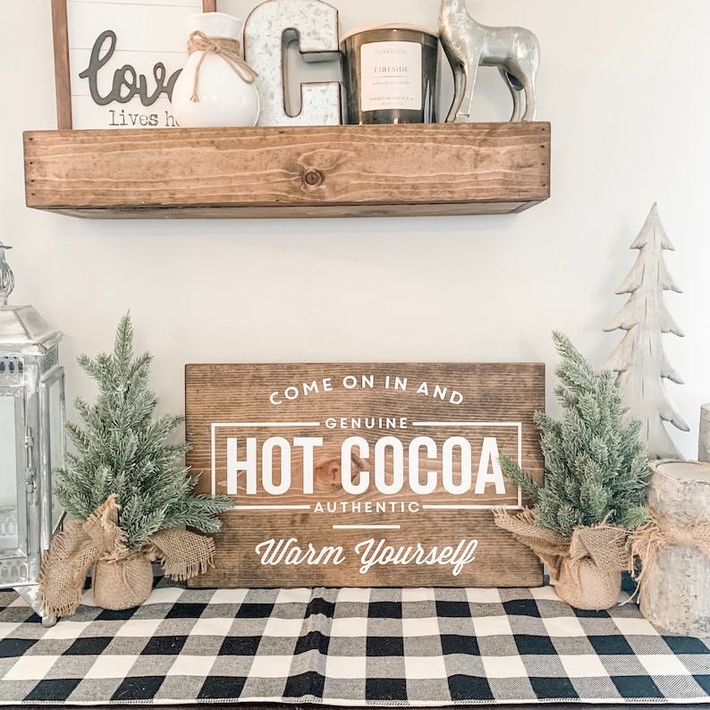 May include: A wooden sign with the text "Come on in and Genuine Hot Cocoa Authentic Warm Yourself" in white lettering. The sign is on a black and white checkered table runner with two small artificial pine trees on either side.