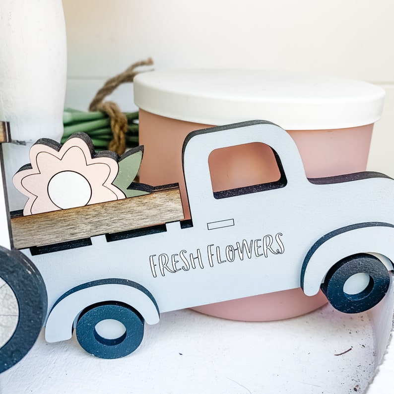 May include: A light blue wooden toy truck with a brown wooden bed, a pink flower, and the words "FRESH FLOWERS" printed on the side.