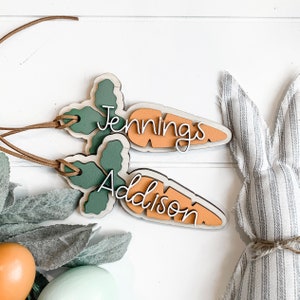 K&ouml;nnte beinhalten: Zwei h&ouml;lzerne Karotten-Anh&auml;nger mit gr&uuml;nen Bl&auml;ttern. Die Karotten sind orange lackiert und die Bl&auml;tter sind gr&uuml;n. Die Karotten tragen die Namen "Jennings" und "Addison" in wei&szlig;.