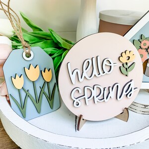 May include: A pink wooden sign with the text "hello spring" in white script. The sign has a small wooden stand and a yellow flower with green leaves attached to the side.