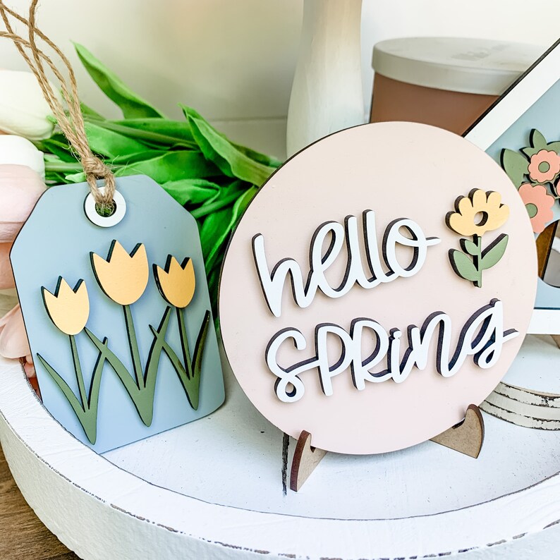 May include: A pink wooden sign with the text "hello spring" in white script. The sign has a small wooden stand and a yellow flower with green leaves attached to the side.