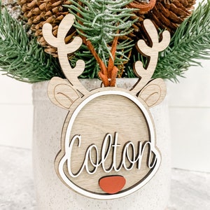 May include: Wooden reindeer ornament with a white outline and the name Colton written in cursive. The ornament has a red nose and is sitting in a white planter with brown pine branches.