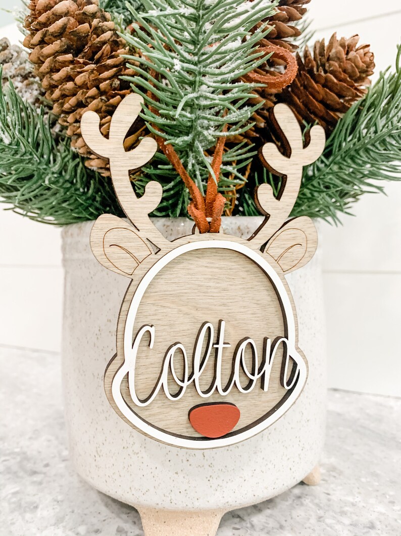 May include: Wooden reindeer ornament with a white outline and the name Colton written in cursive. The ornament has a red nose and is sitting in a white planter with brown pine branches.