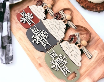 May include: Three wooden Christmas ornaments shaped like mugs with names and snowflakes. The top ornament is red and says "CRISWELLS". The middle ornament is black and says "GRANDKIDS". The bottom ornament is green and says "THE MILLERS".