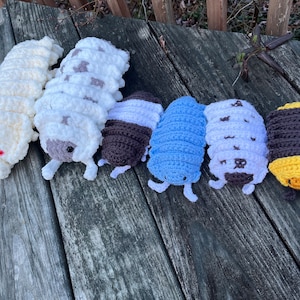May include: Six crocheted pill bugs in various colours, including white, brown, blue, and yellow. The pill bugs are arranged in a row on a wooden surface.