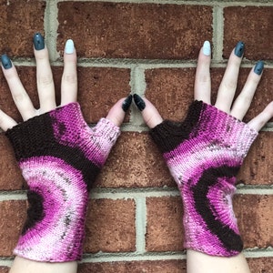 May include: A pair of fingerless gloves with a pink and brown ombre pattern. The gloves are knitted and have a loose fit.