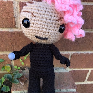 Puede incluir: Muñeca amigurumi de crochet con un mohawk rosa, vestida con un atuendo negro y sosteniendo una pequeña bola gris.